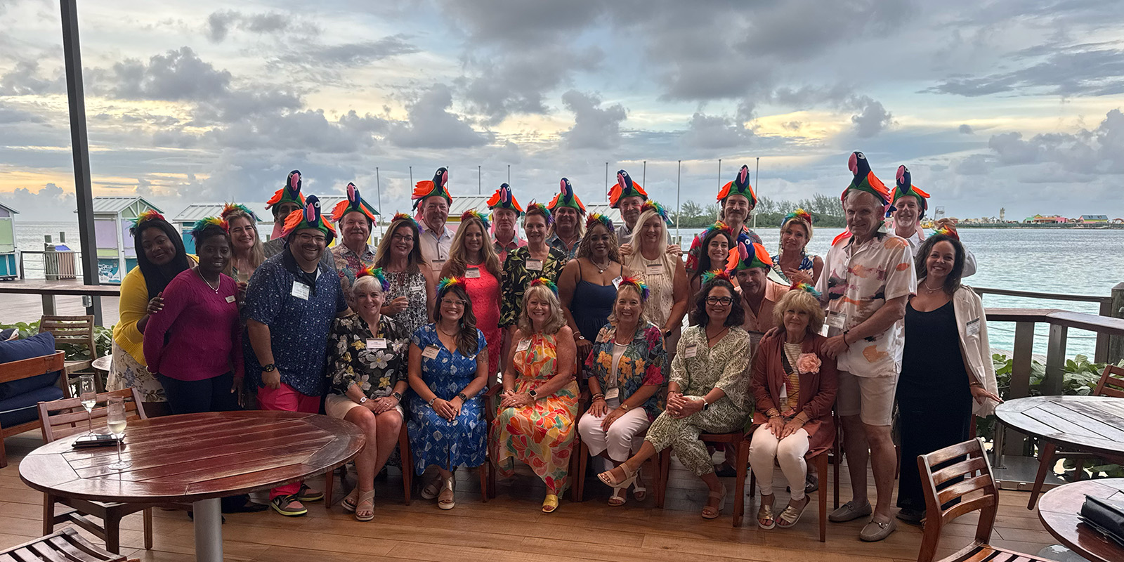 Council members and guests in matching parrot hats, gathered on a waterfront deck at dusk.