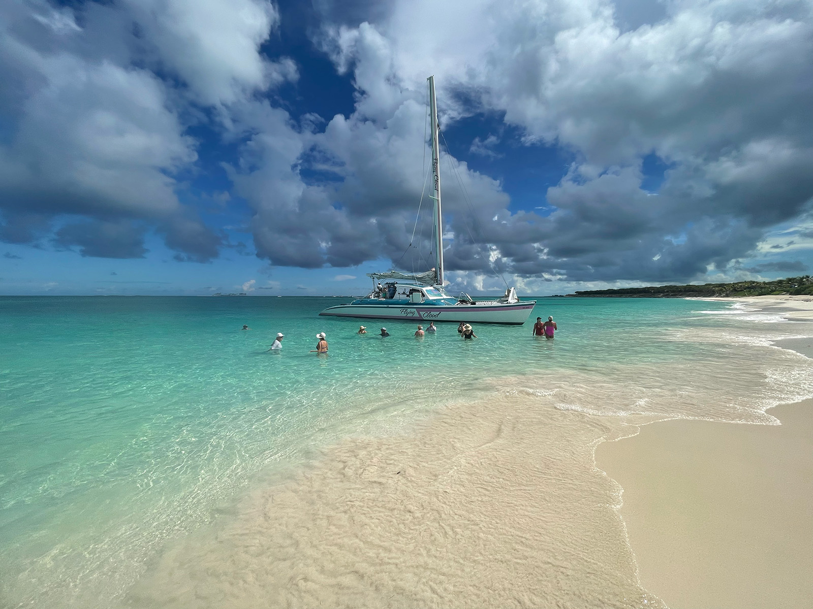 A sailing catamaran anchored off a white-sand beach in clear turquoise water, with guests wading ashore.